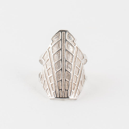 A silver-colored ring with a geometric pattern, featuring a large face lifting design, placed against a white background.